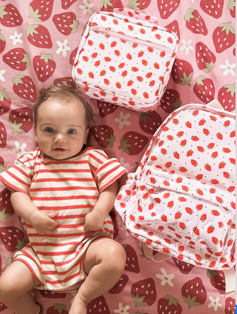 baby girl laying down with a strawberry fields mini backpack and a lunch bag